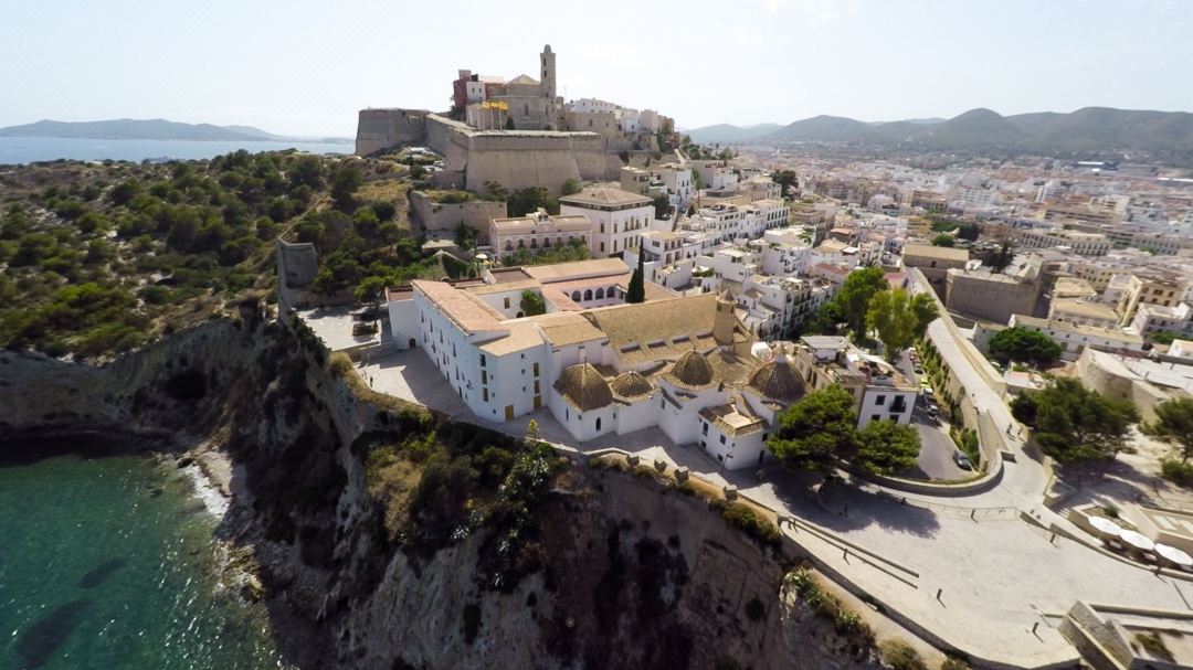 A bedroom at Mirador de Dalt Vila featuring a green onyx bathroom visible through glass doors, with a Juliet balcony overlooking the Ibiza harbor.