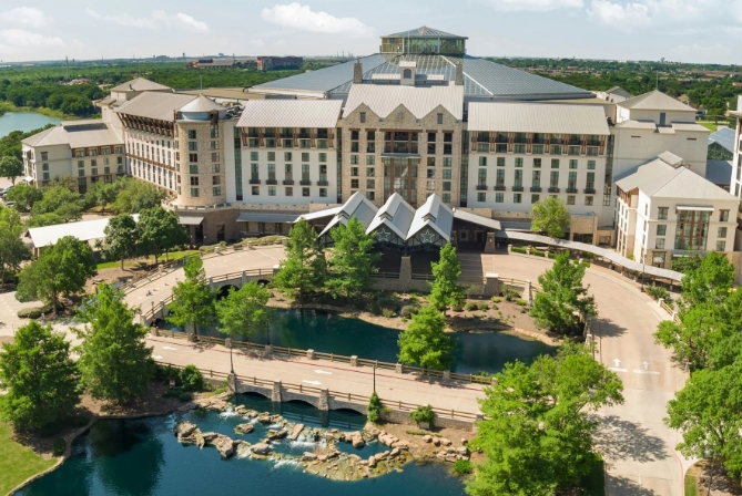 A massive glass atrium filled with lush indoor gardens, a winding river replica, and rustic Texas-style limestone buildings under a soaring skylight roof.
